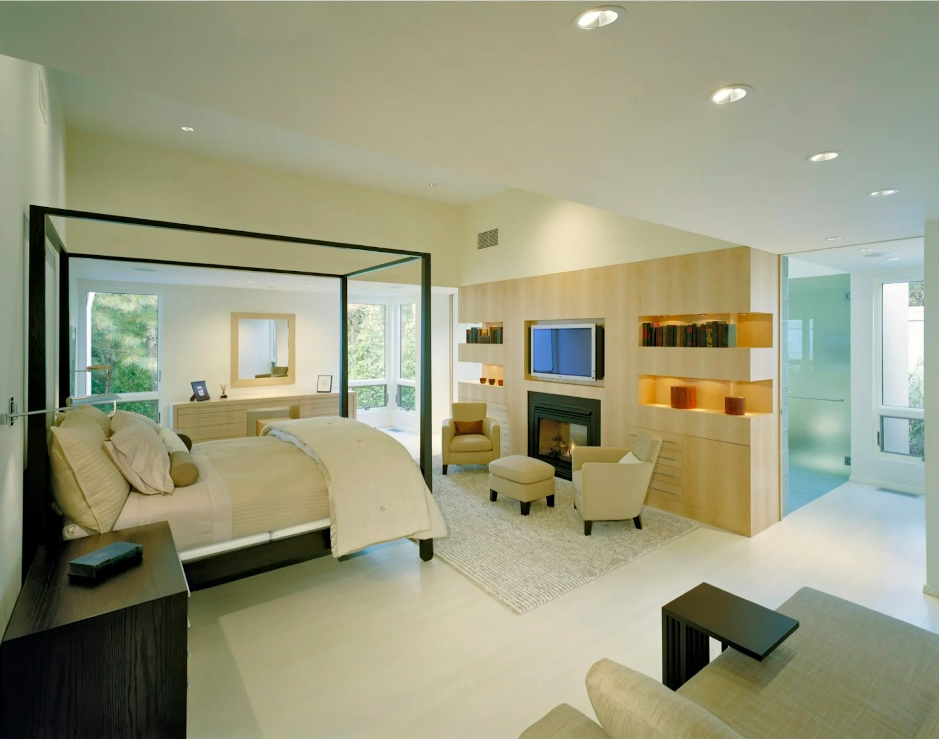 Luxurious modern master bedroom suite with black canopy bed, fireplace, built-in shelving, and spa-like bathroom visible through glass partition by Lisa Dubin Architect.
