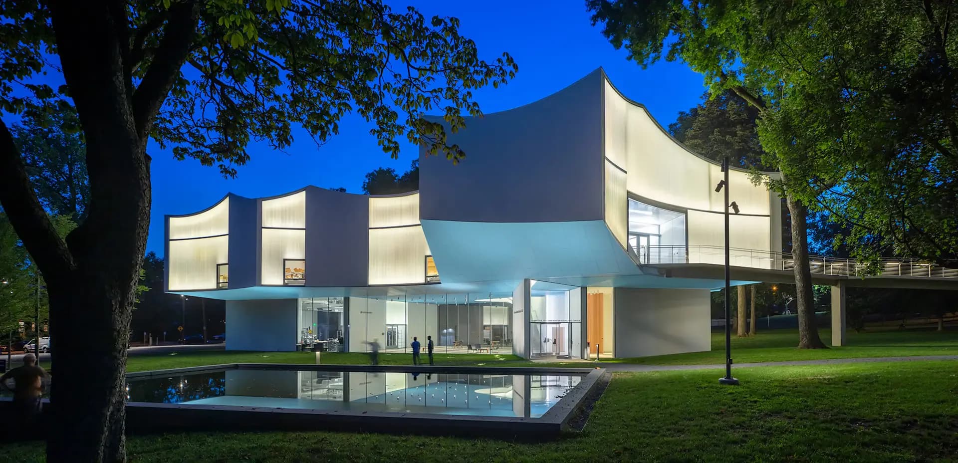 Modern residential architecture with geometric white and blue facades, illuminated at dusk, featuring a reflecting pool and manicured lawn by L'Observatoire International.