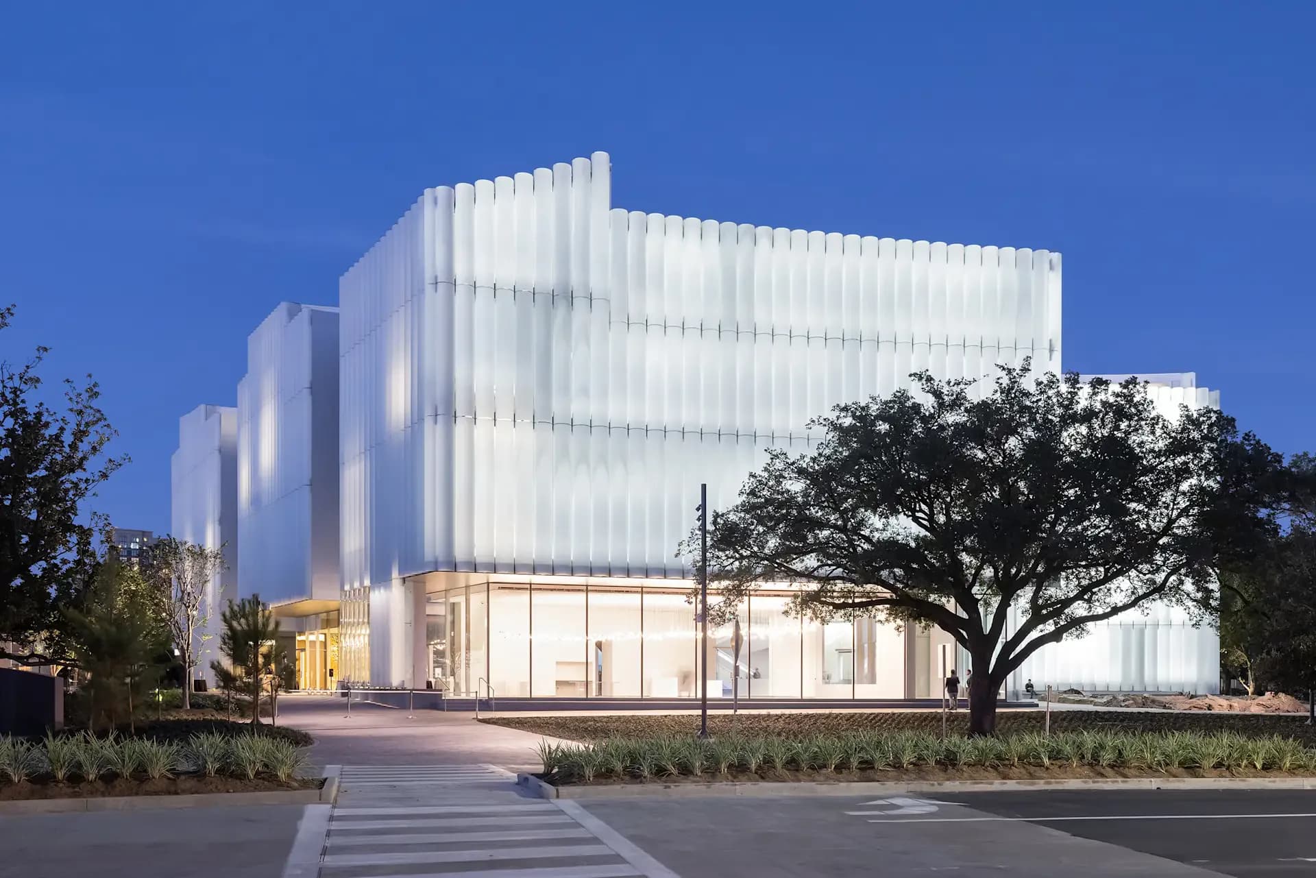 Modern white translucent facade building with vertical ribbed panels, evening-lit ground floor retail spaces, and manicured landscaping by L'Observatoire International.