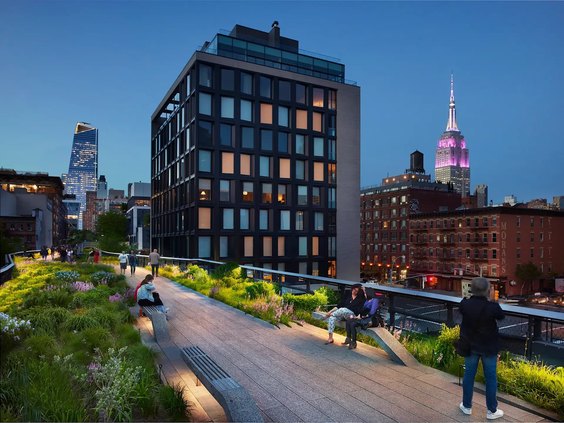 The High Line elevated park in New York features a modern dark-facade residential building alongside landscaped pathways with native plantings and pedestrians, with the Empire State Building illuminated in the background, by L'Observatoire International.