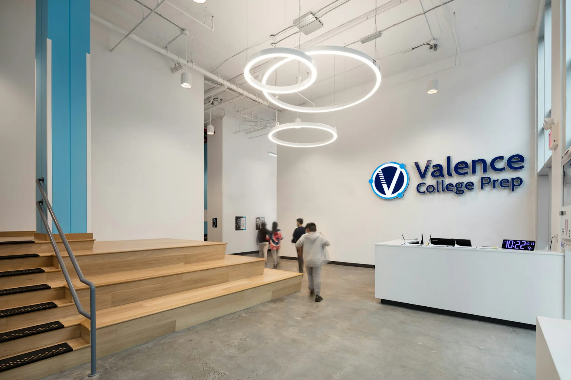 Valence College Prep reception lobby featuring modern circular pendant lighting, wooden stairs, glass partitions, and clean white walls with blue accent elements by Loci Architecture PLLC.