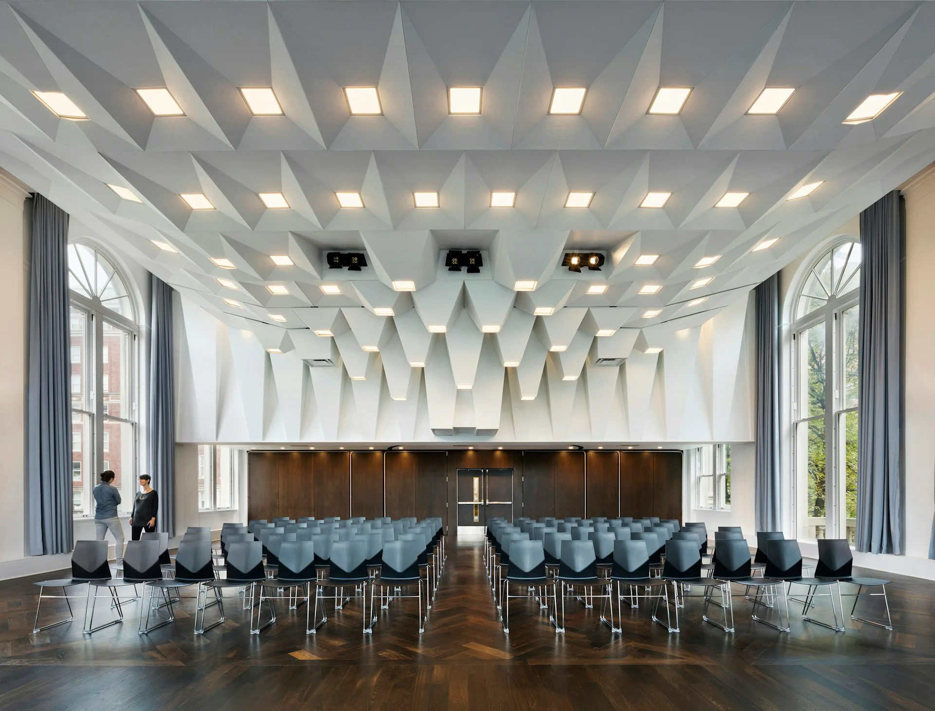 Modern assembly hall with geometric white coffered ceiling, integrated lighting, dark wood stage backdrop, and blue chairs arranged in rows by LTL Architects.
