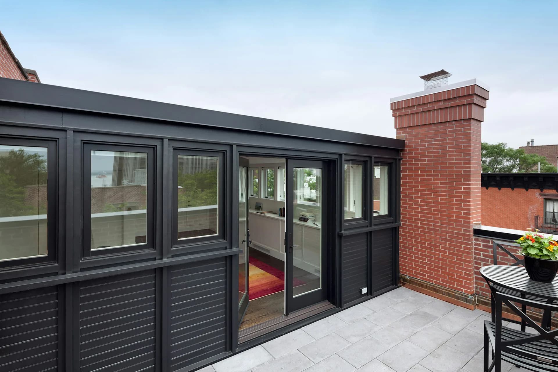 Modern black-framed glass pavilion with louvered lower panels adjoining a brick chimney on a paved rooftop terrace by LUBRANO CIAVARRA ARCHITECTS.