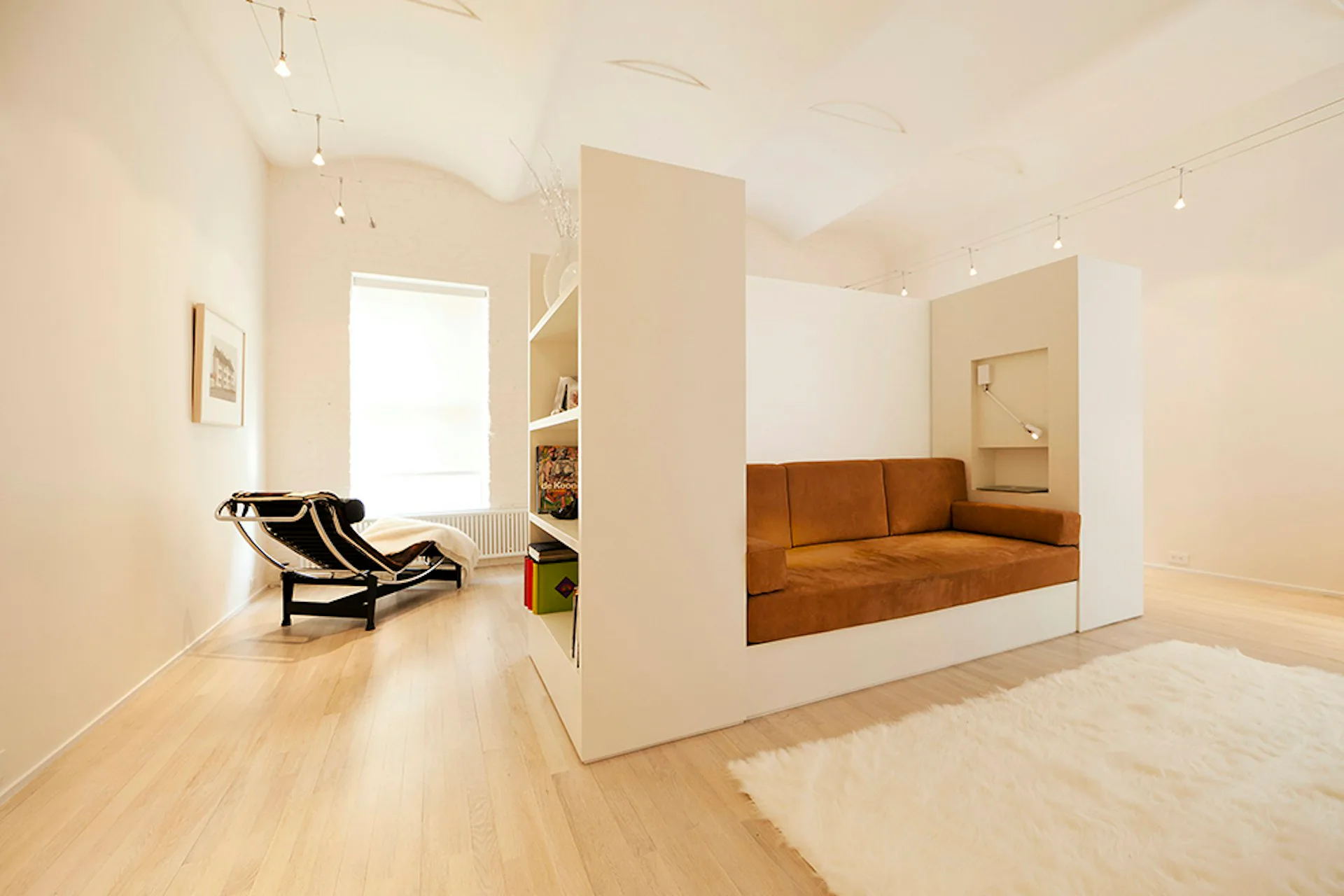 Modern minimalist studio apartment with cream walls, light wood flooring, and a central white partition featuring a burnt-orange upholstered bed nook, designed by LUBRANO CIAVARRA ARCHITECTS.