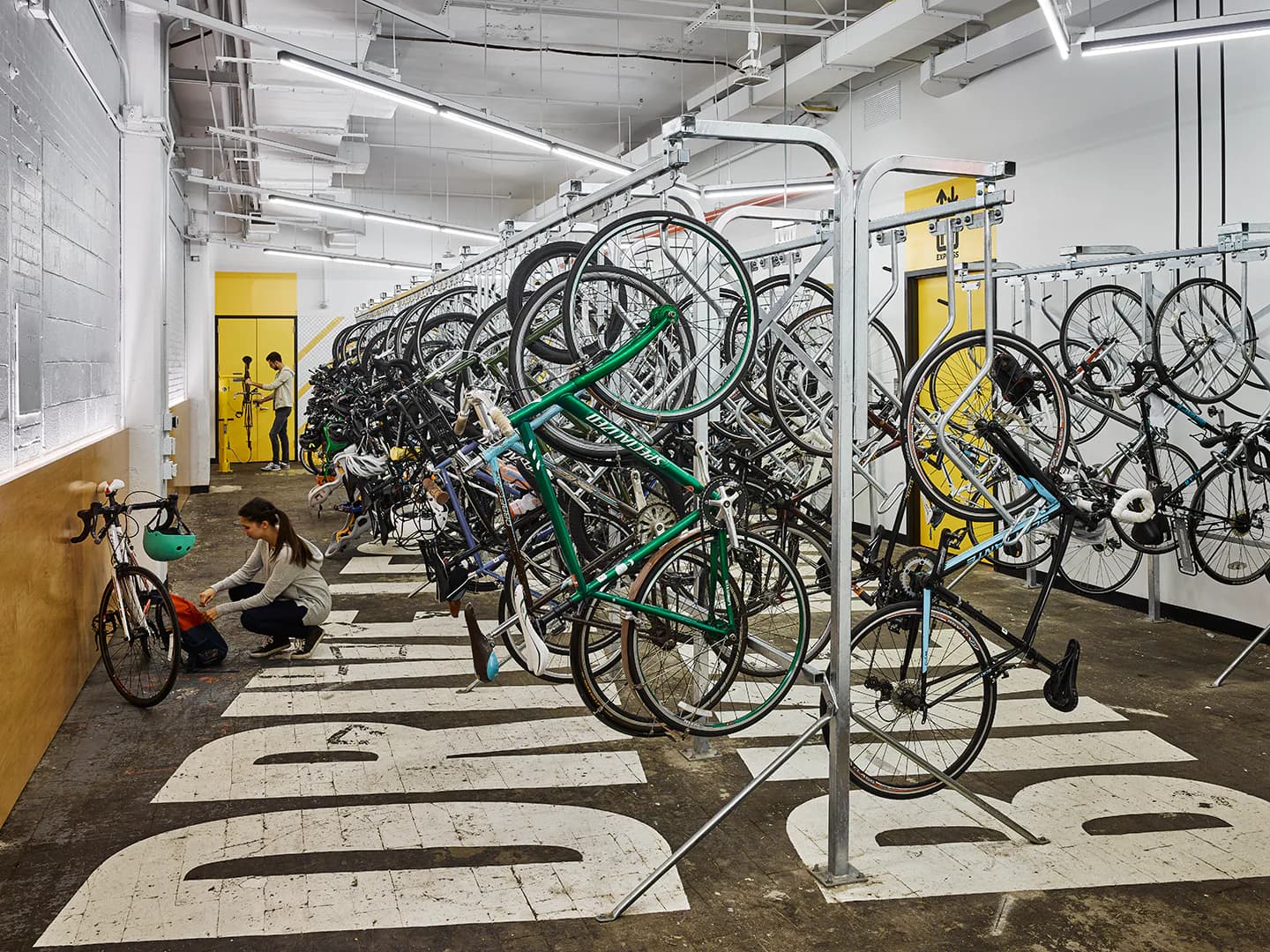 Modern bike storage facility with rows of bicycles on metal racks, concrete floor with markings, yellow accents, and industrial ceiling by Lumen Architecture.