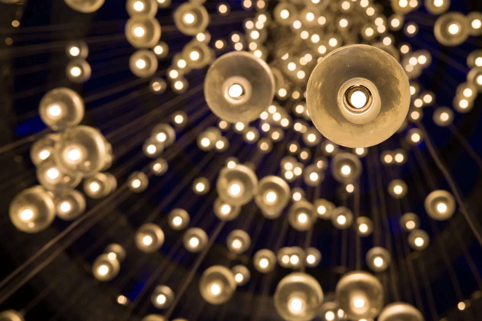 Overhead view of a dramatic chandelier installation featuring numerous warm-toned pendant lights arranged in a radiating pattern against a dark ceiling by Lux Foundry.