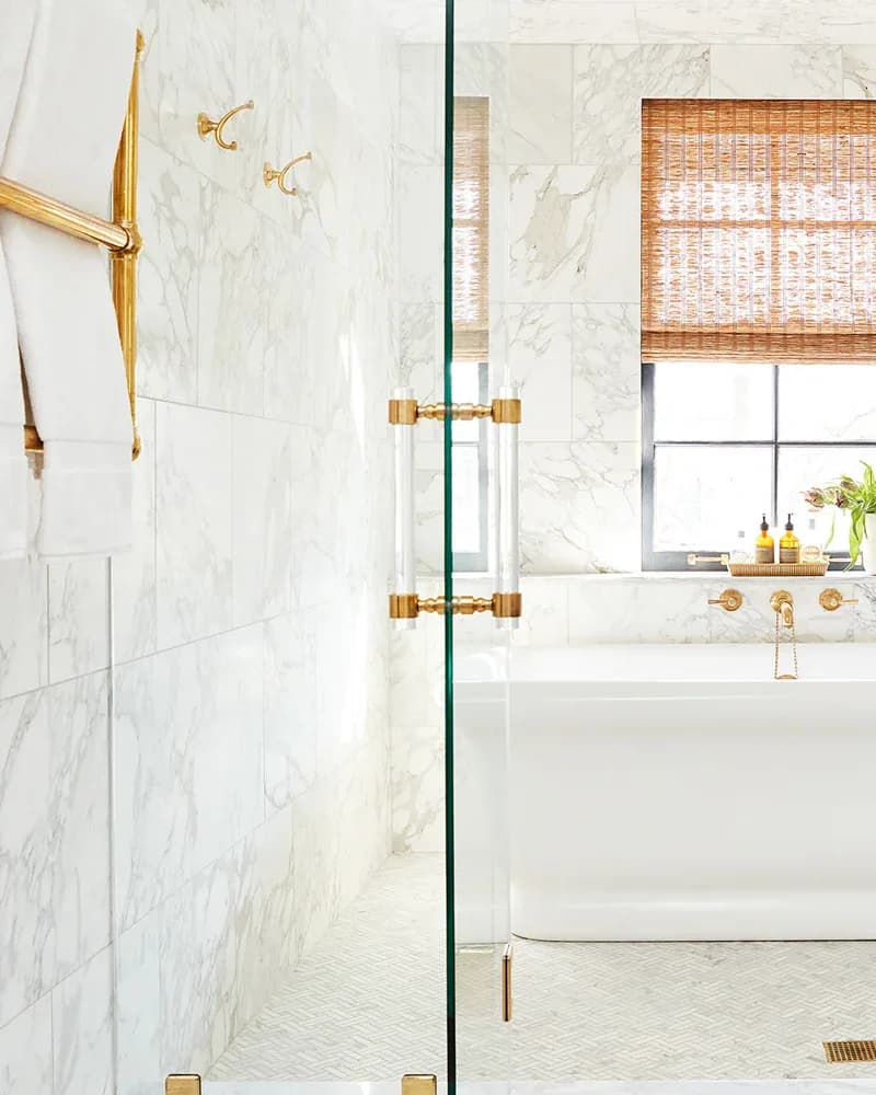 Luxurious marble bathroom with brass fixtures, glass shower enclosure, and woven window shade by LuxHoldUps.