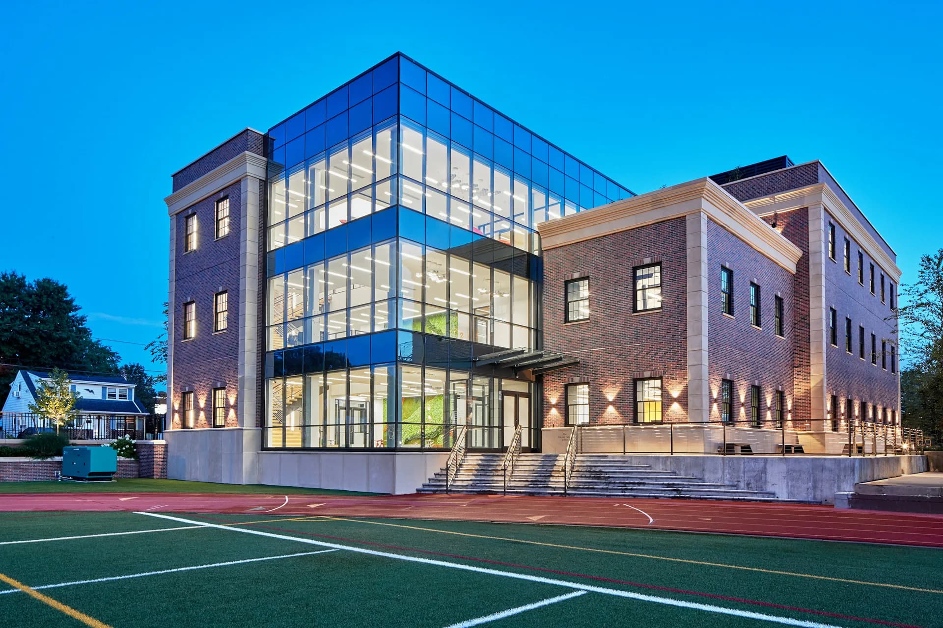 Modern institutional building with illuminated glass curtain wall facade adjoining brick structures, overlooking a red and green sports court by Mancini Duffy.