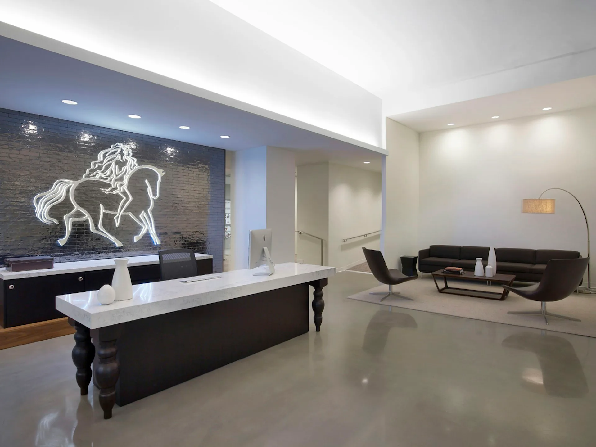 Modern commercial lobby featuring dark polished concrete floors, white reception desk, neon horse artwork, and contemporary black seating area designed by Mancini Duffy.