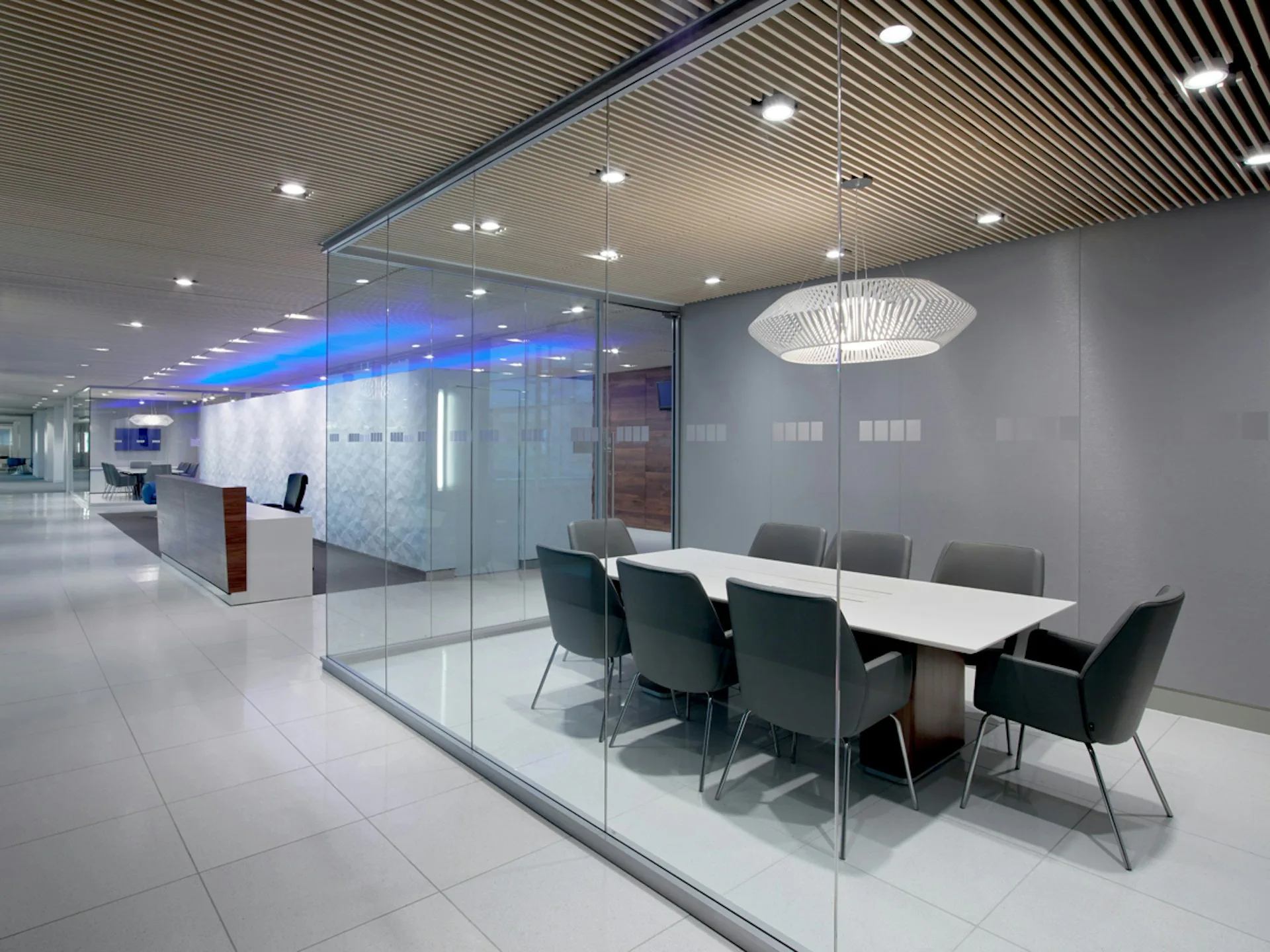 Modern office meeting room with glass partitions, white table, dark chairs, blue accent lighting, and linear ceiling details by Mancini Duffy.