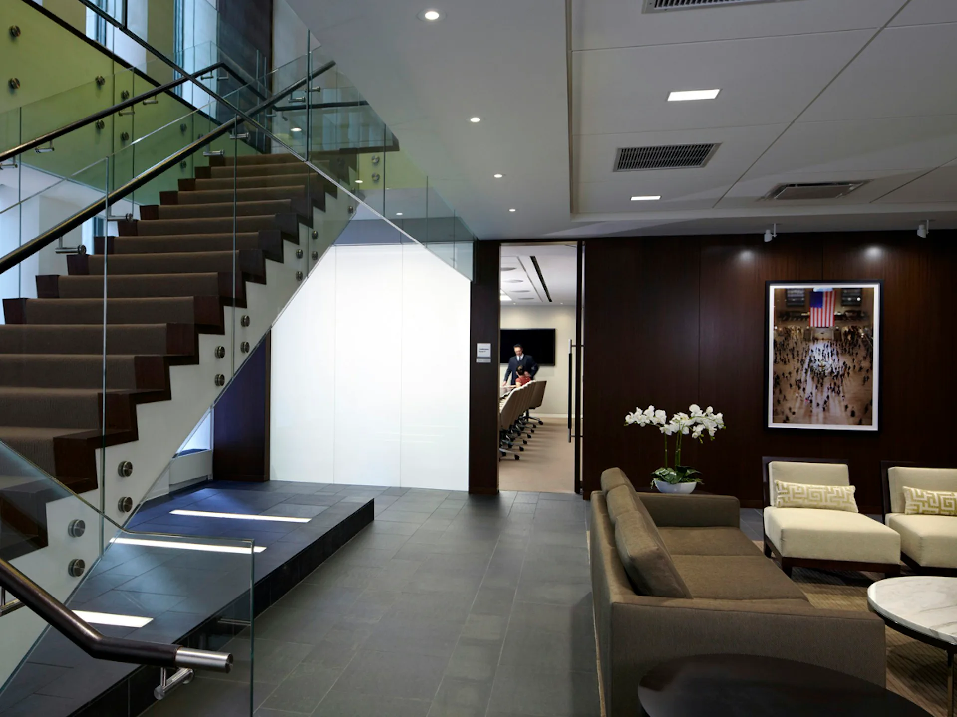 Modern lounge with dark accent wall, cream seating, wood staircase with glass railings, and minimalist lighting by Mancini Duffy.