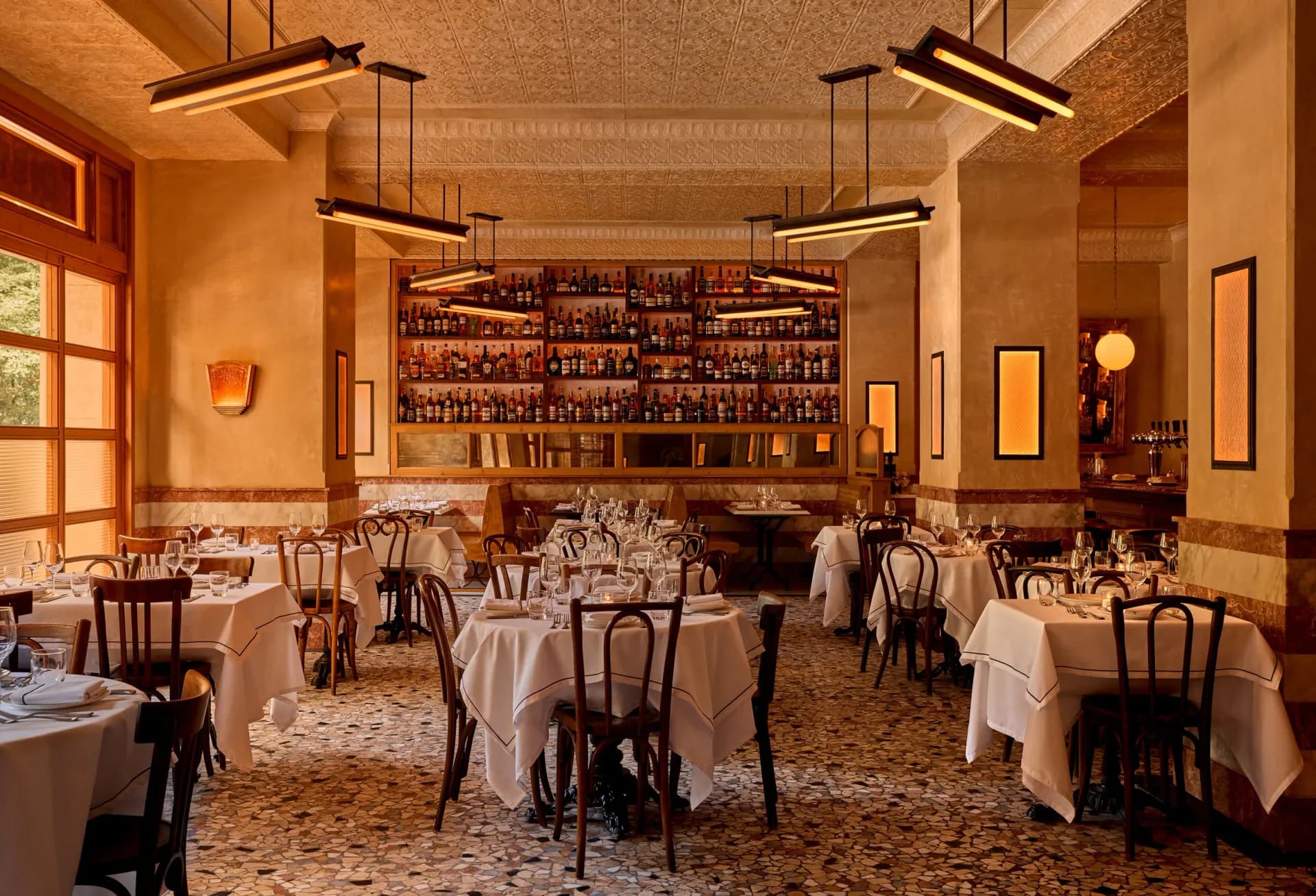 Elegant fine dining restaurant interior with warm terracotta walls, white tablecloths, bentwood chairs, and a backlit bar displaying bottles, designed by Map Design Studio.