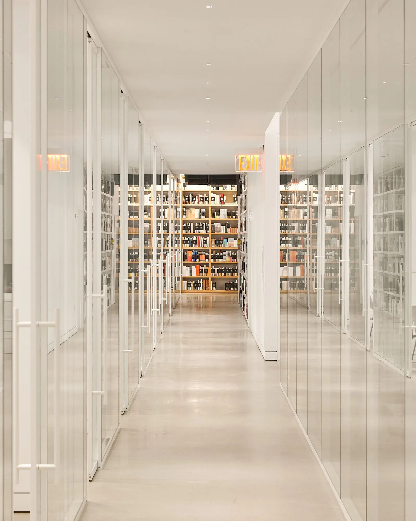 A pristine library corridor with white glass-fronted cabinetry lining both sides, warm wood shelving visible beyond, and polished light flooring, designed by Map Design Studio.