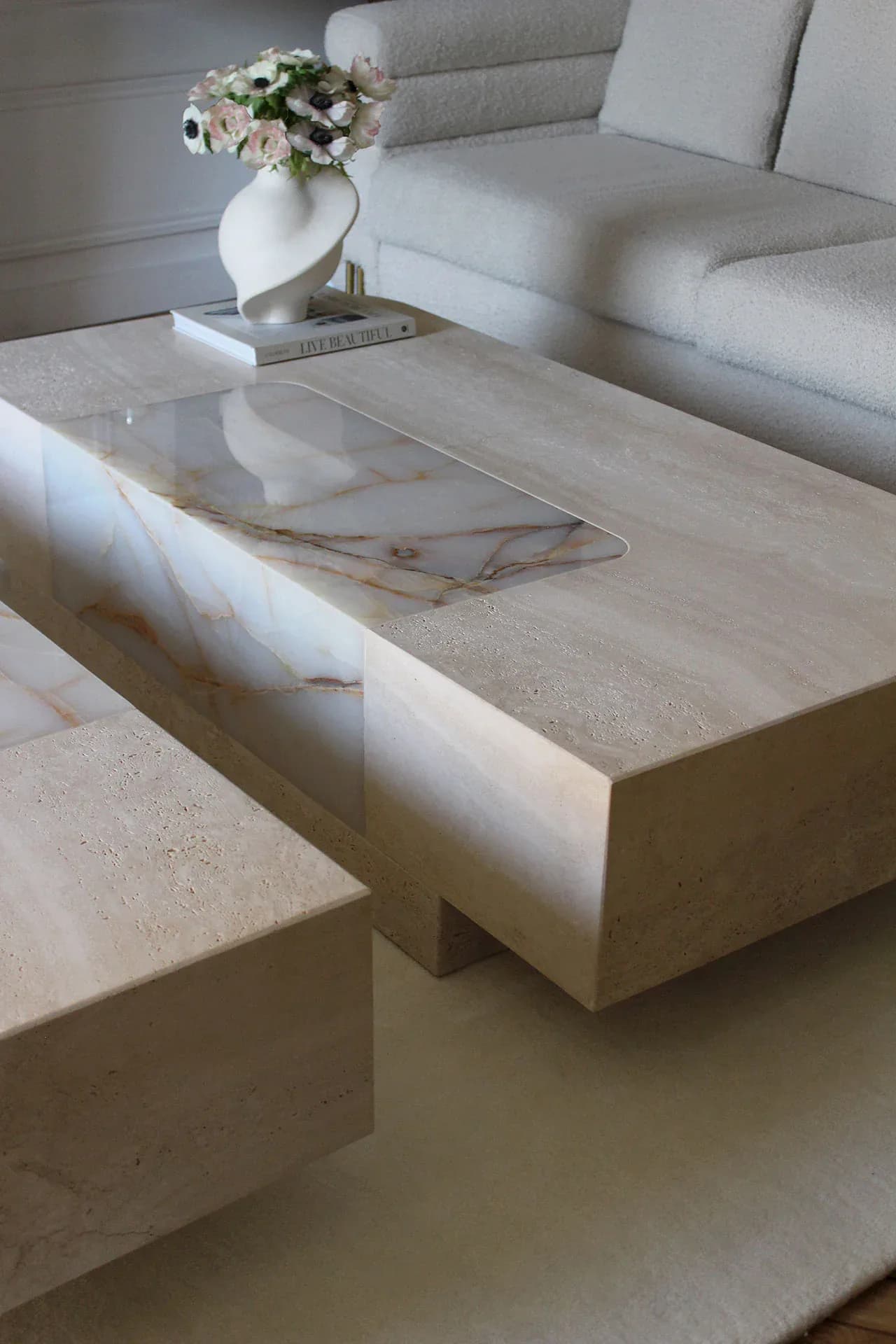 Minimalist living room with cream travertine coffee table and matching side table, white marble inlay detail, soft gray upholstered sofa, and white ceramic vase with flowers by Marbera.