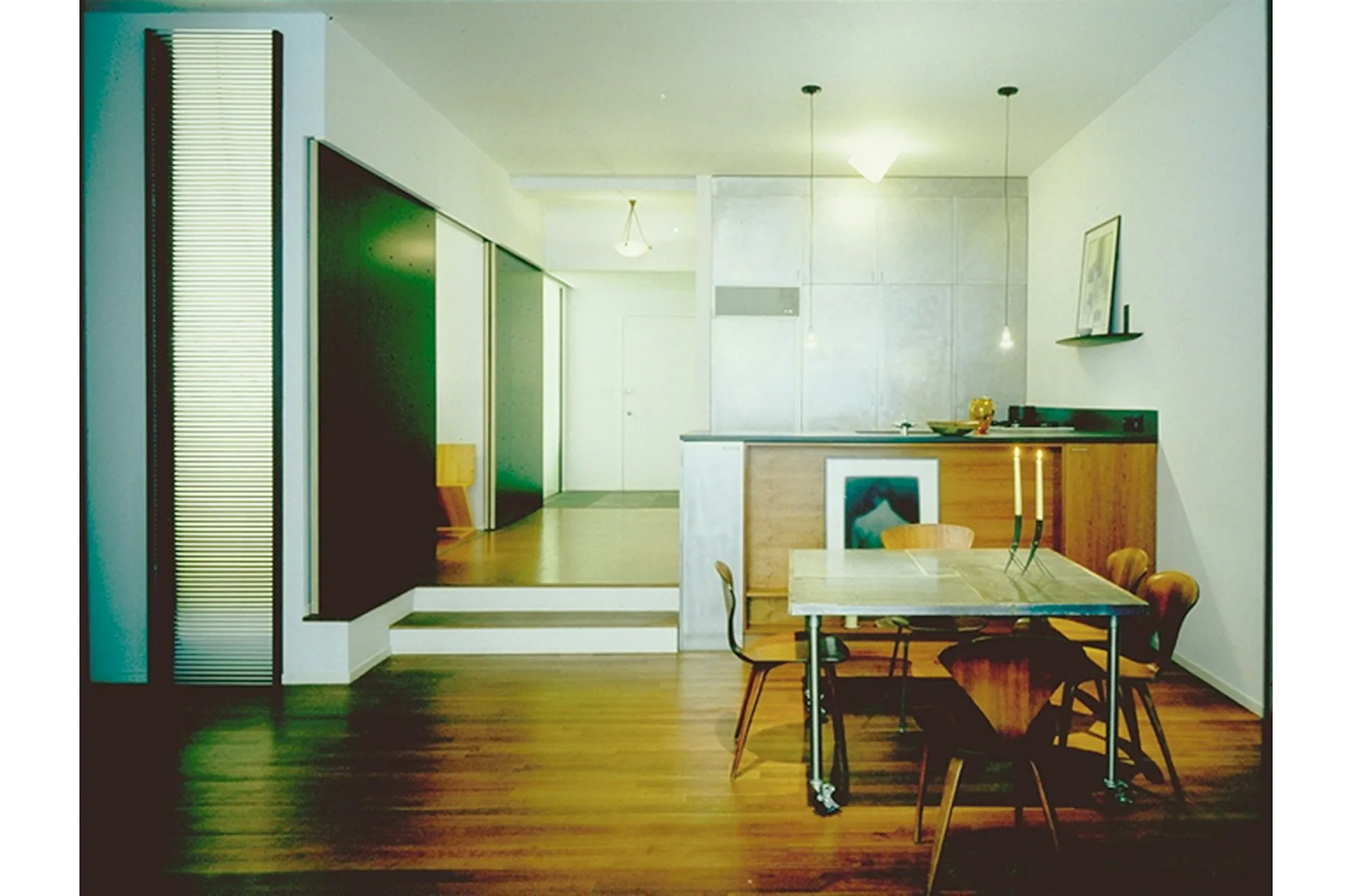Modern open-plan loft interior with warm wood flooring, dining table with mid-century chairs, kitchen cabinetry, and glass partition walls defining spaces by Marble Fairbanks.