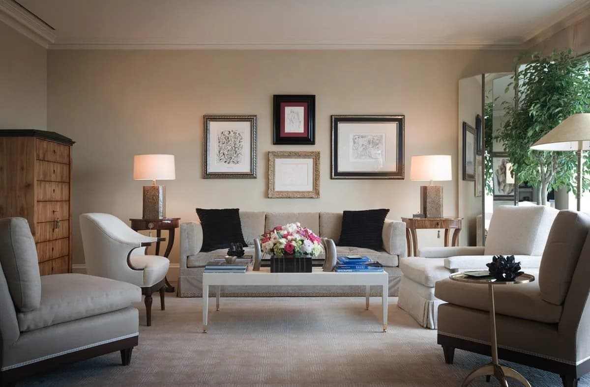 Sophisticated living room with paired gray upholstered chairs, white coffee table, framed artwork, table lamps, and wood cabinetry by Mark Hampton LLC.