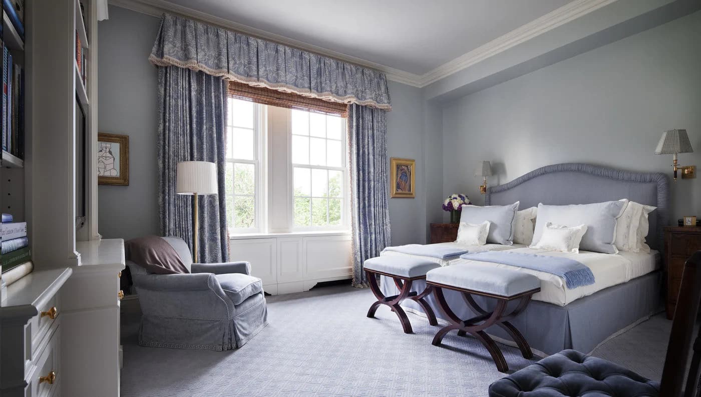Serene bedroom with blue-gray walls, layered window treatments, upholstered bed with white linens, and coordinated furnishings by Mark Hampton LLC.