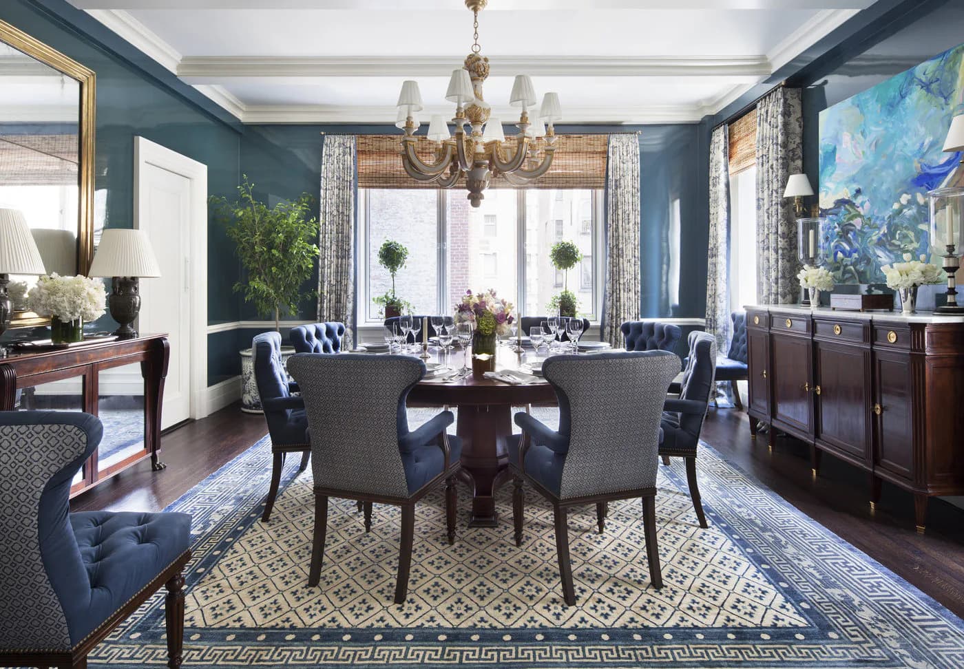 Formal dining room with teal walls, round mahogany table, upholstered chairs, brass chandelier, patterned area rug, and layered drapery by Mark Hampton LLC.