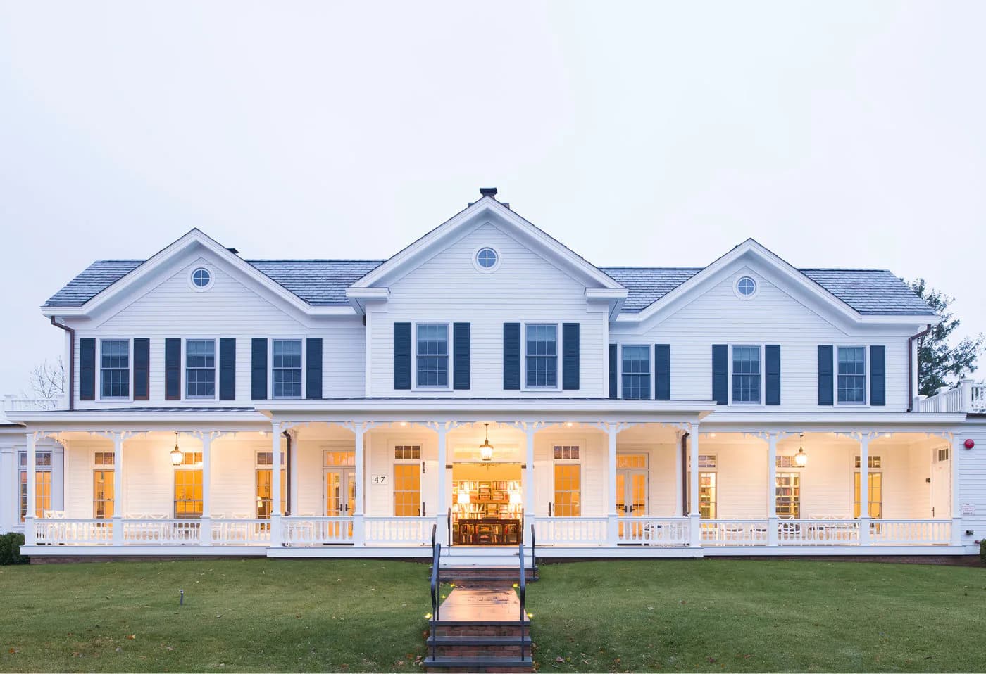White farmhouse exterior with blue shutters, covered porch with columns, illuminated at dusk, expansive lawn and manicured grounds by Mark Hampton LLC.