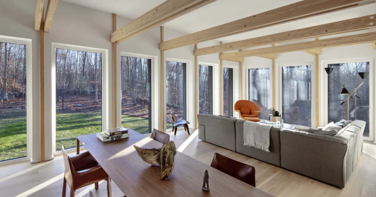 Sunlit modern sunroom with expansive windows overlooking wooded landscape, featuring gray upholstered seating, wooden ceiling beams, and minimalist furnishings by Marner + Marner.