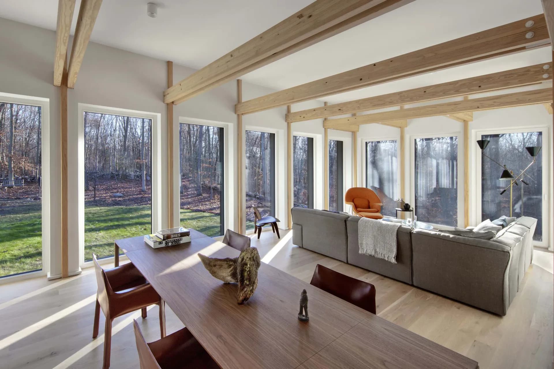 Sunlit open-concept living and dining space with expansive windows, warm wood beam ceiling, gray upholstered seating, and natural views by Marner + Marner.