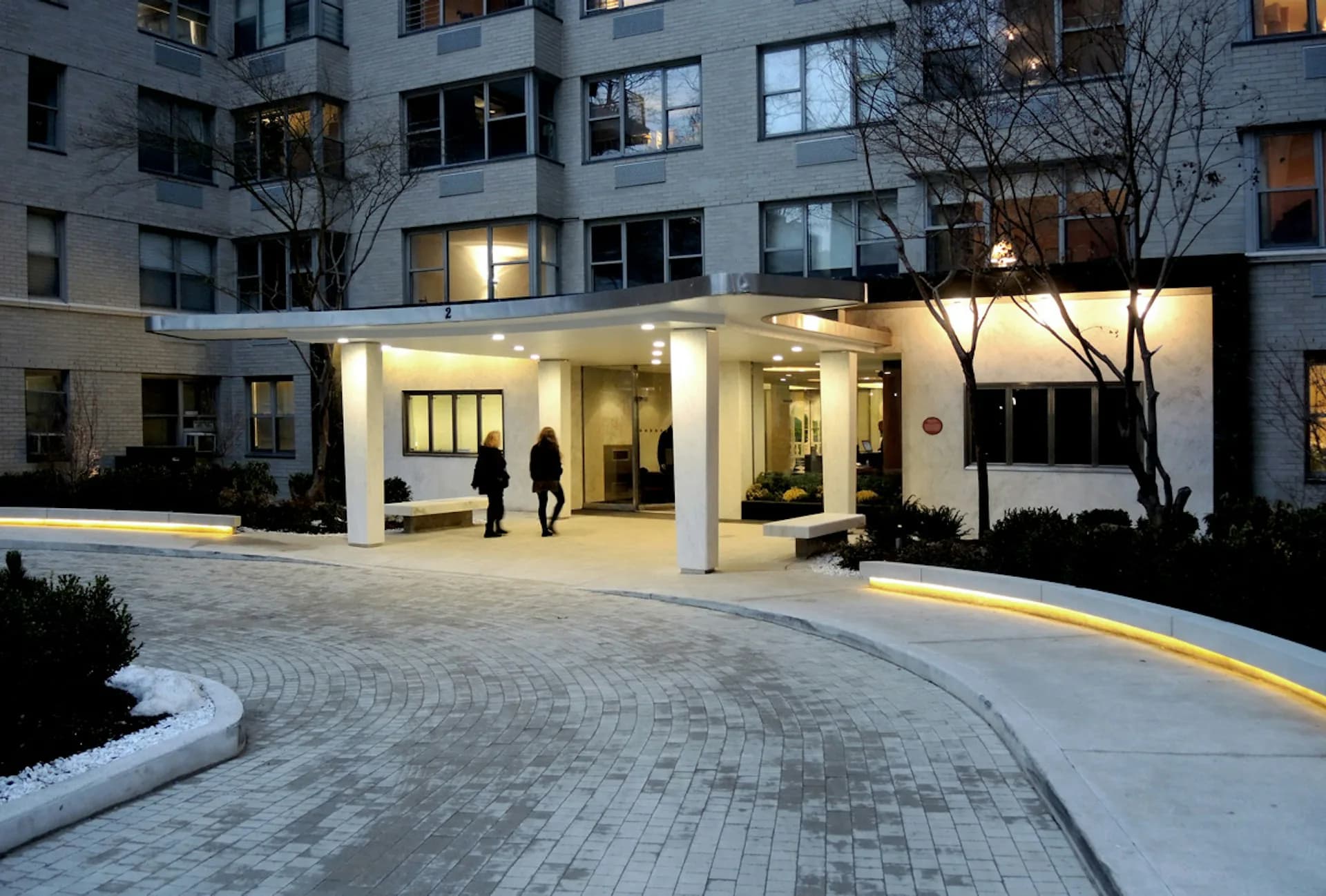 Modern building entrance with illuminated white canopy, paved courtyard with curved pattern, and lit facade designed by Marpillero Pollak Architects.