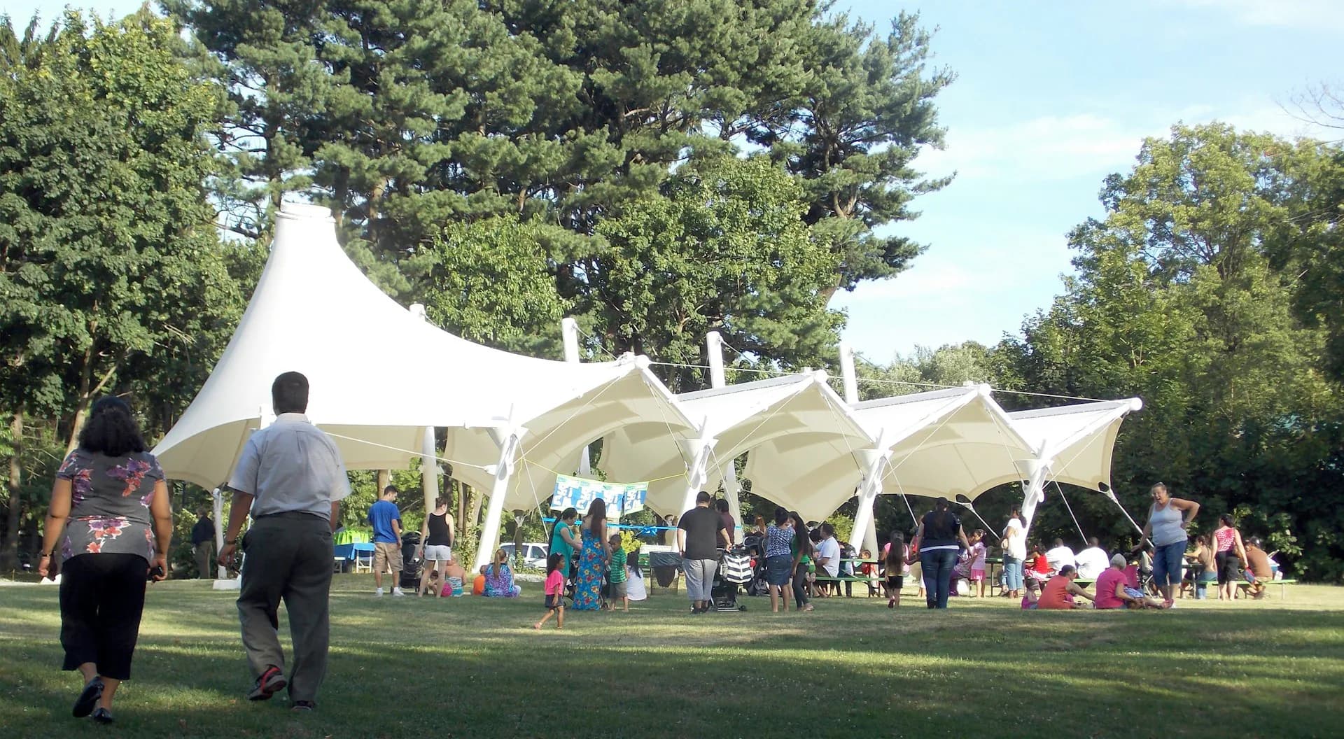 White tensile fabric pavilions with multiple peaks installed in a park setting surrounded by trees, with visitors gathered for an outdoor event, designed by Marpillero Pollak Architects.