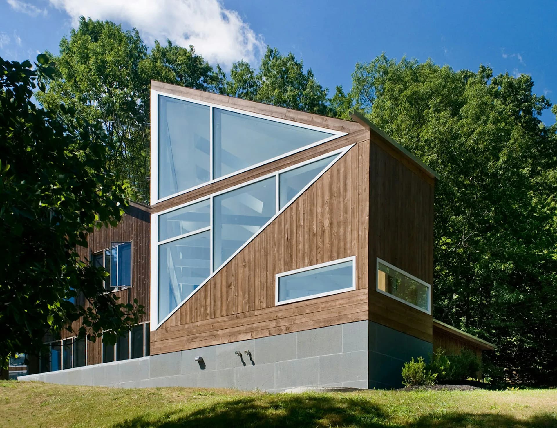 Modern residential building with angled wooden cladding, large triangular windows, and concrete base set against green landscape by martin holub architects.