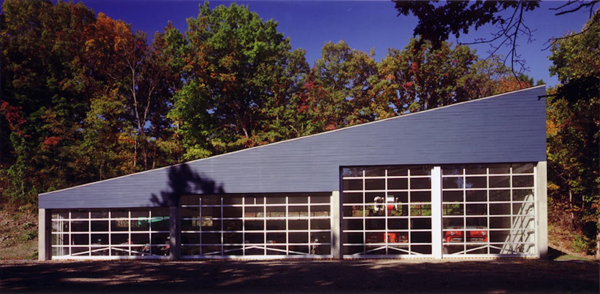 Modern white concrete building with expansive grid-pattern glass garage doors set among autumn trees, designed by Martin Holub Architects.