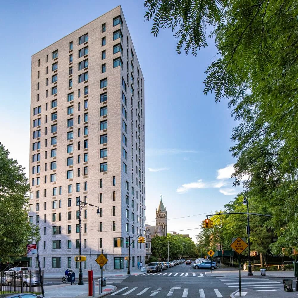 Tall residential apartment building with tan brick facade and regular window grid, positioned on a tree-lined urban street corner with a church steeple visible in the background by Marvel.