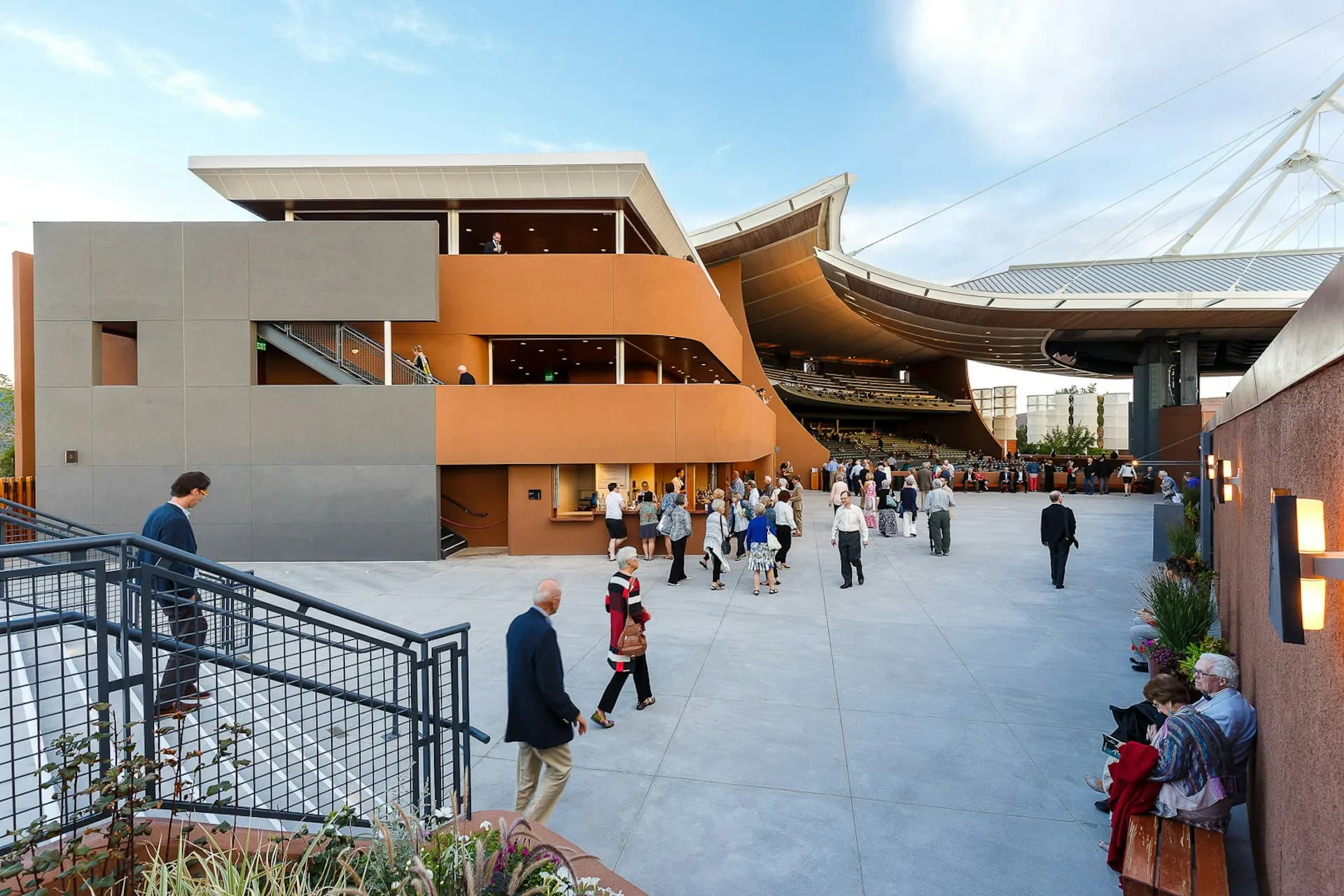 Modern institutional courtyard with white and orange angular building, curved wooden architectural elements, and multiple visitors in a paved plaza by Matiz Architecture & Design.