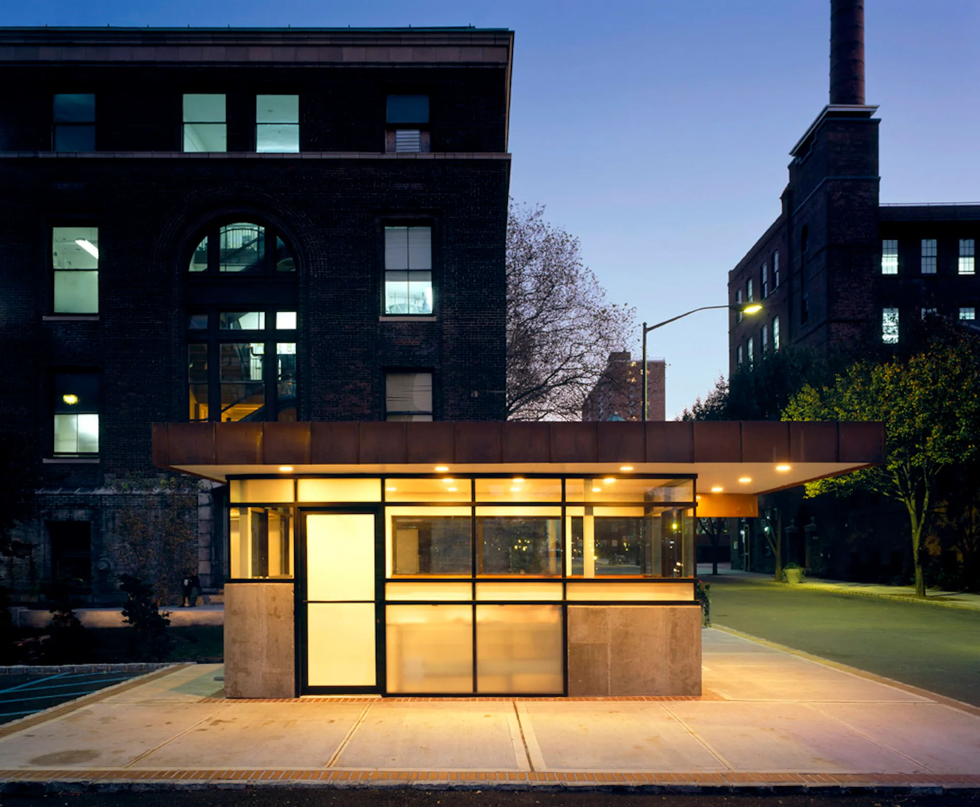 Modern glass and metal pavilion structure with warm interior lighting, situated in front of historic brick buildings at dusk by Matiz Architecture & Design.