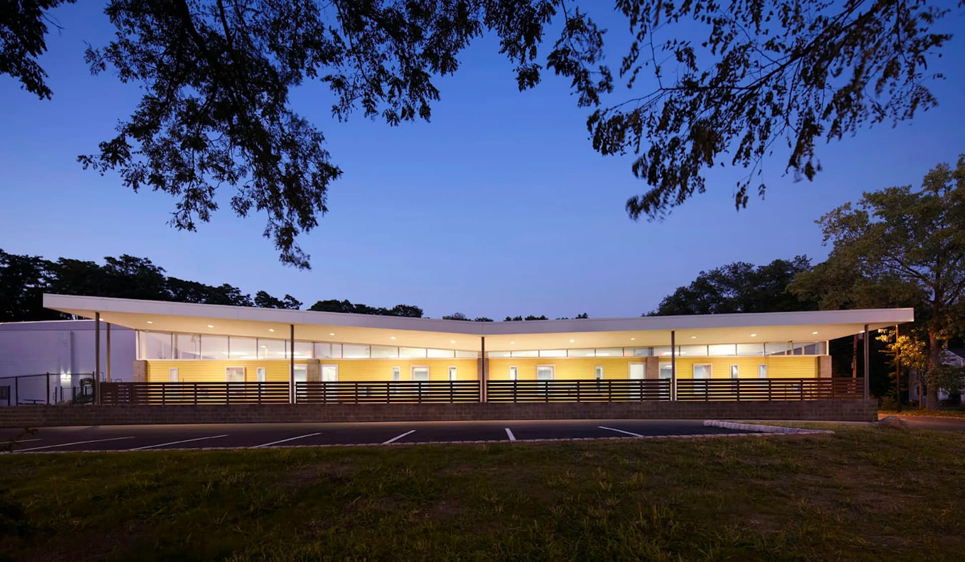 A modern single-story institutional building with extensive glazing illuminated at dusk, horizontal linear design with metal fencing, set in a landscaped grounds by Matiz Architecture & Design.