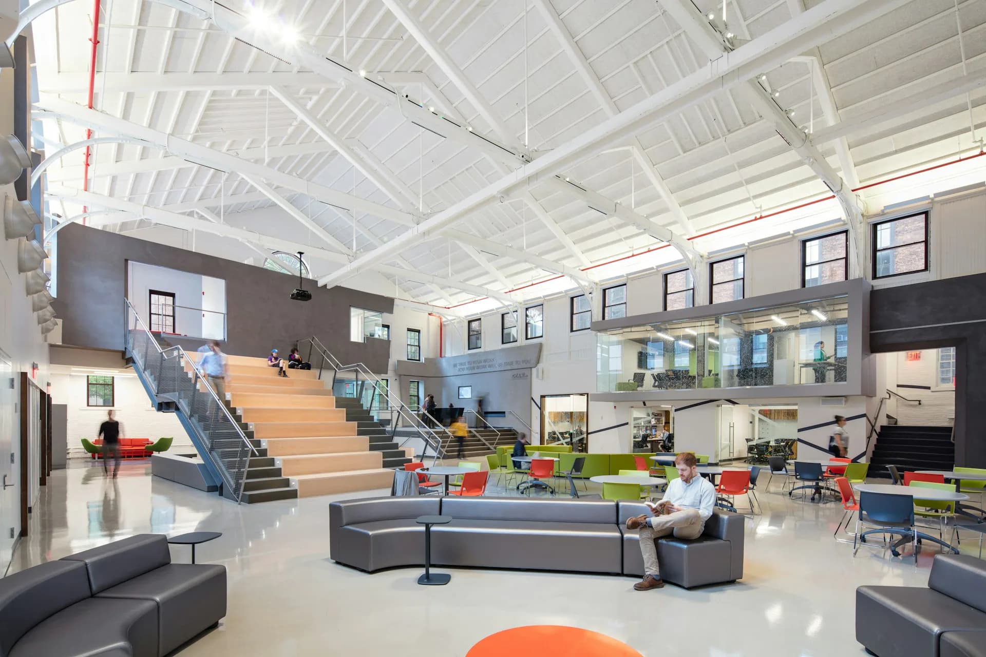 Modern school or community center atrium with double-height timber staircase, polished concrete floors, abundant natural light from clerestory windows, colorful furniture clusters, and industrial ceiling details by Matiz Architecture & Design.