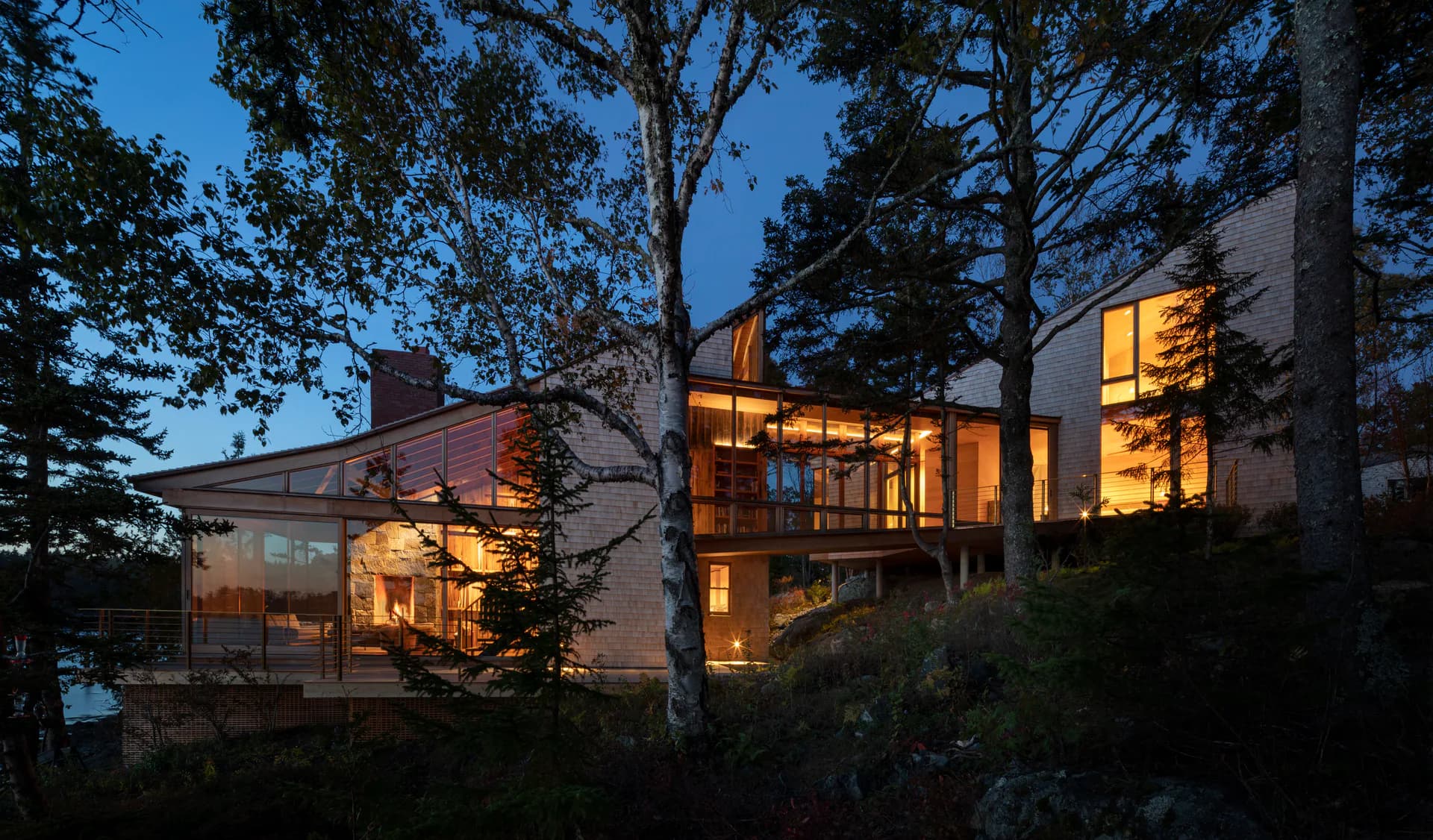 Modern residential house photographed at dusk with warm interior lighting, featuring geometric forms, extensive glazing, and natural wood framing among mature trees by Matthew Baird Architects.