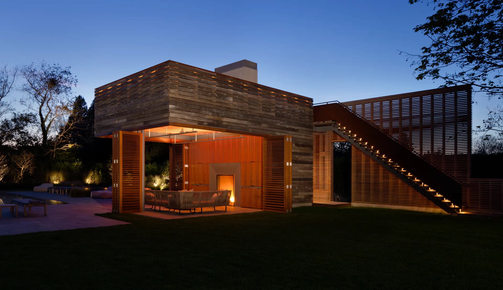 Modern wooden residential structure illuminated at dusk with warm interior lighting, featuring horizontal slat detailing and angular roofline by Matthew Baird Architects.