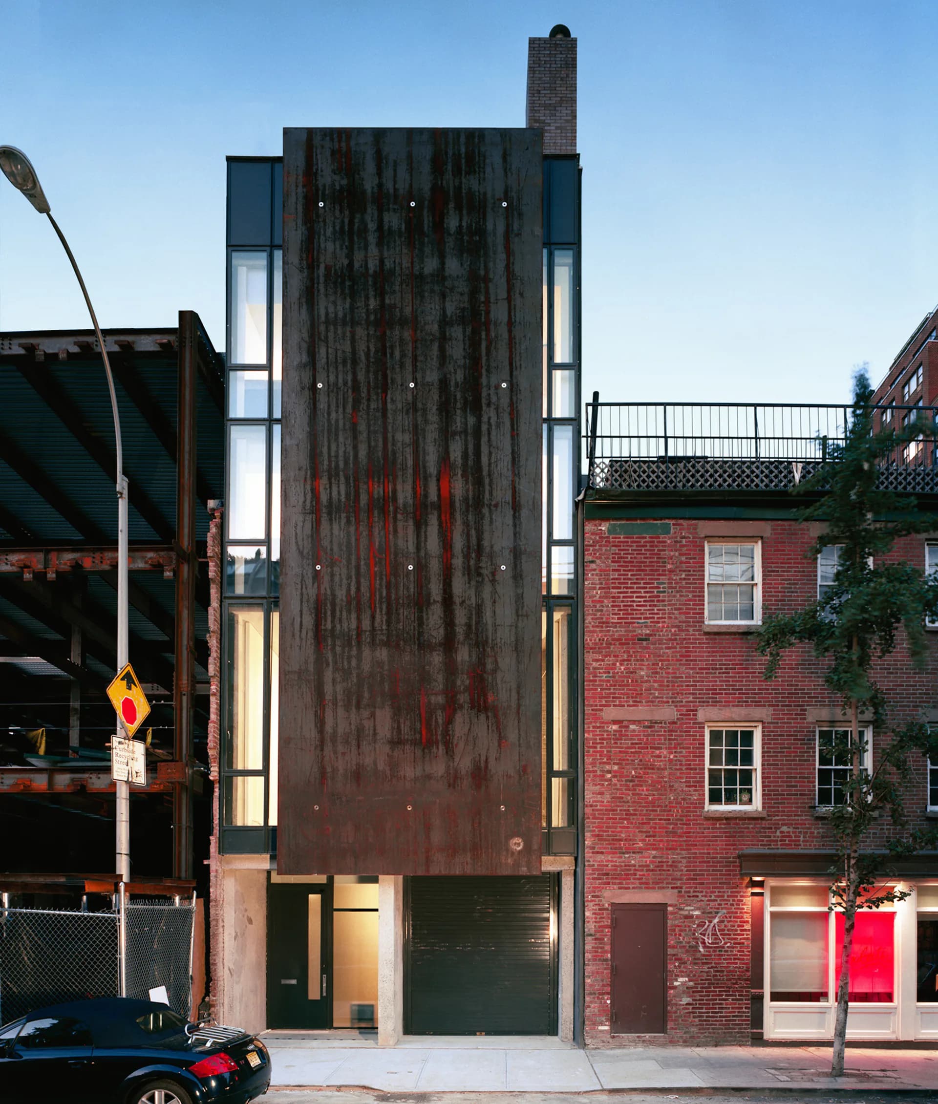 Narrow dark timber-clad residential building with illuminated side windows and chimney, flanked by brick structures on an urban street by Matthew Baird Architects.