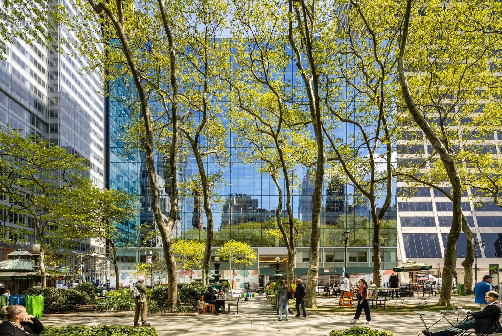 Urban plaza with mature trees, seating areas, and modern glass buildings, a landscaped public space design by MdeAS Architects.