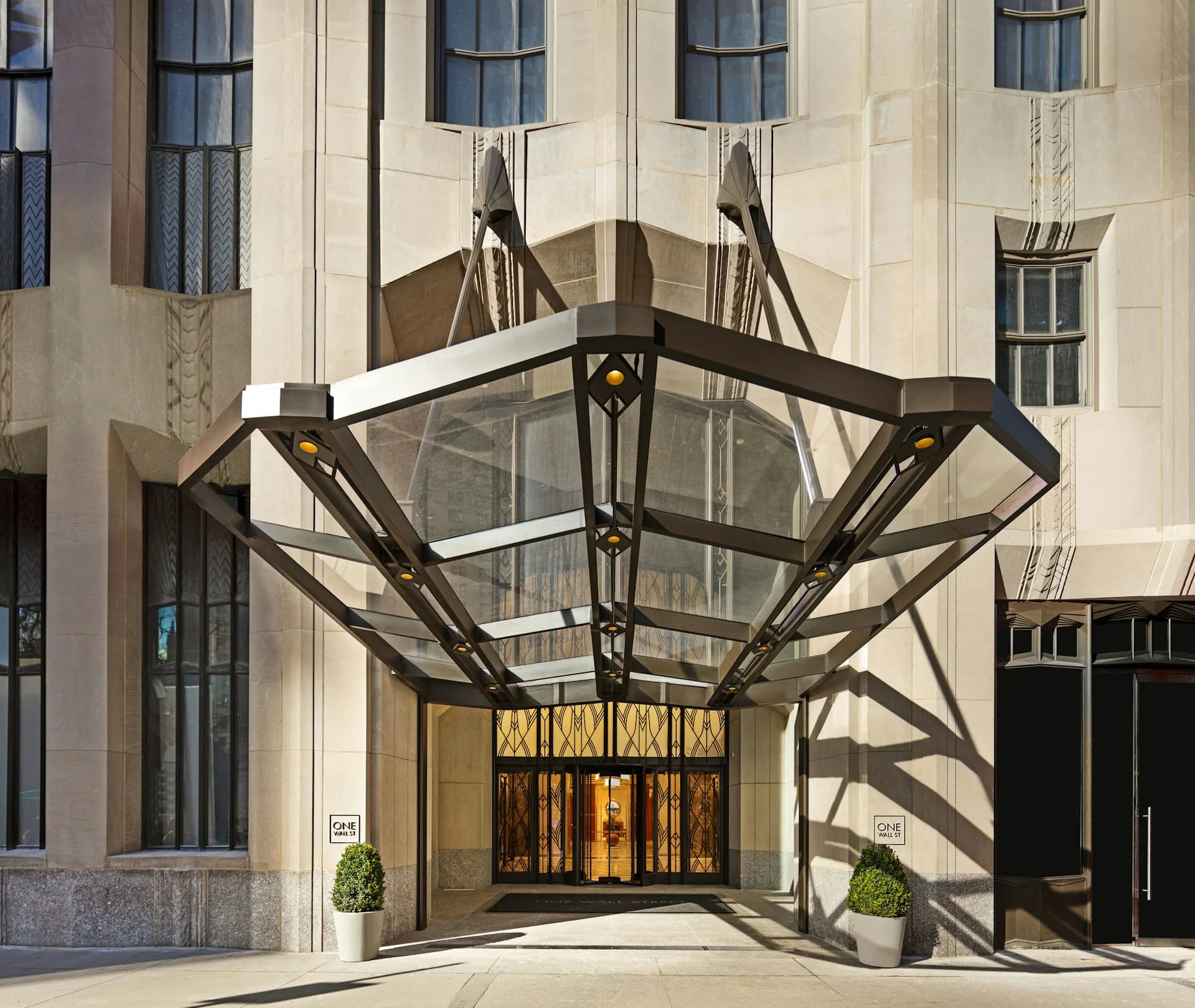Modern bronze canopy entrance with geometric angular design shelters golden doors against cream limestone facade by MdeAS Architects.