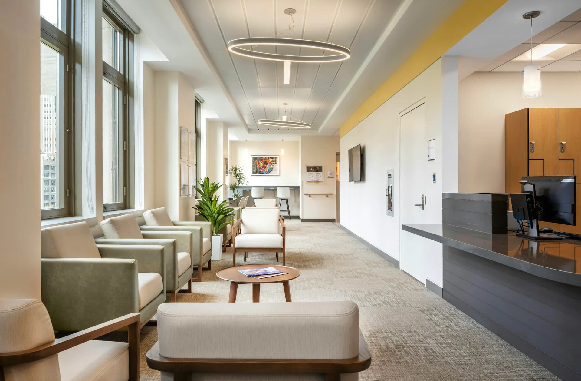 Modern medical office corridor with waiting area featuring green upholstered seating, potted plant, round side table, natural light from floor-to-ceiling windows, and contemporary ceiling fixtures by MDSzerbaty Associates Architecture LLC.