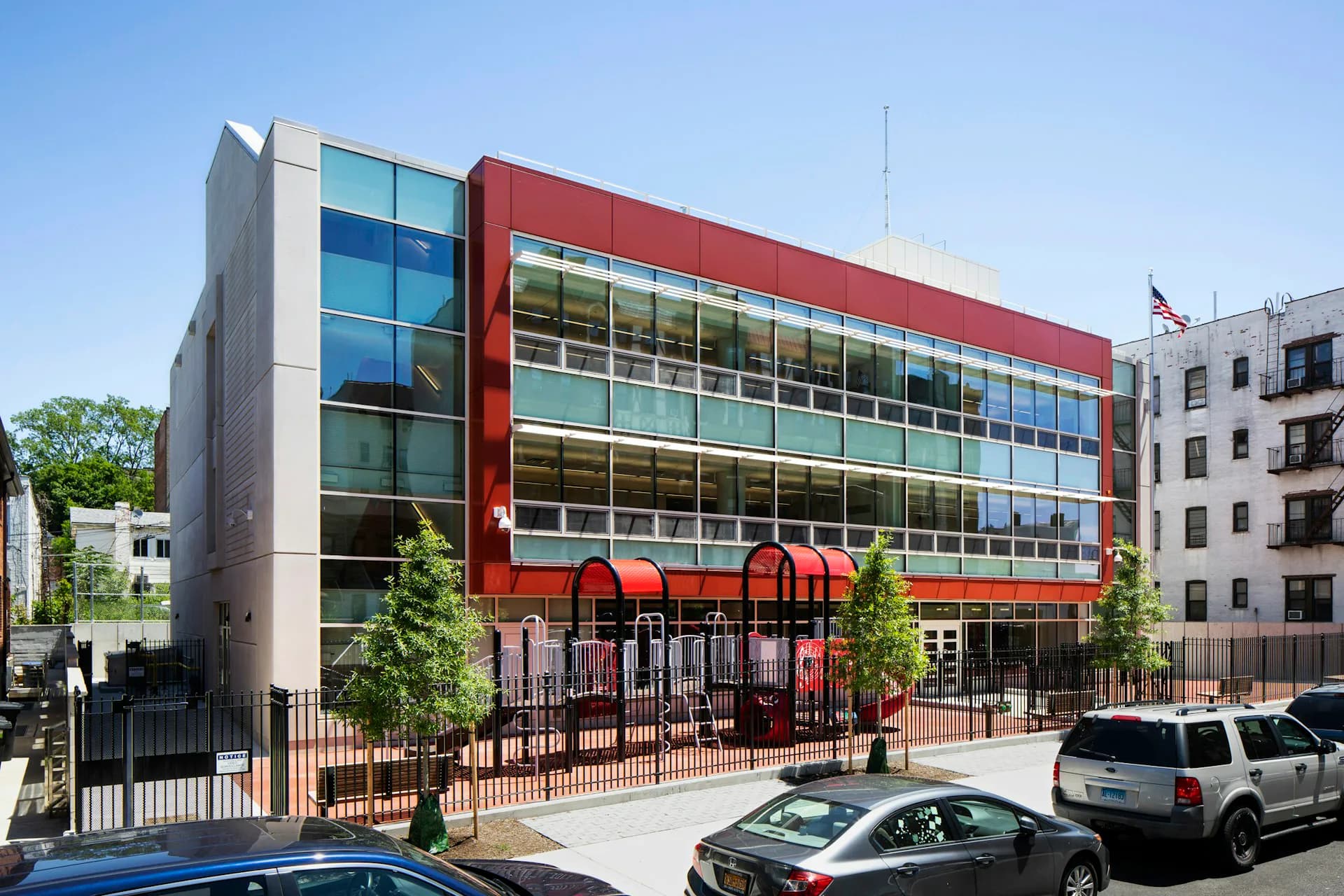 Contemporary mixed-use building with red metal facade, glass windows, and ground-level retail/community space with brick courtyard by MDSzerbaty Associates Architecture LLC.