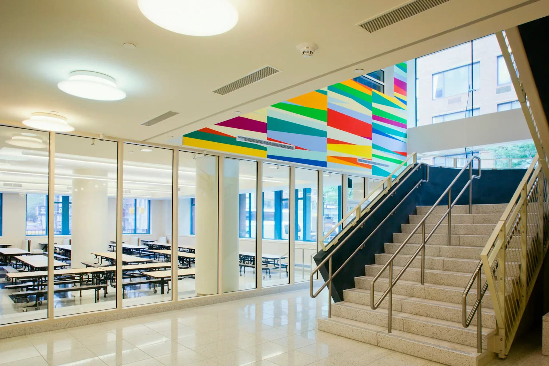 Modern institutional hallway featuring colorful suspended geometric ceiling installation, glass-fronted offices, and marble floors with staircase, designed by MDSzerbaty Associates Architecture LLC.