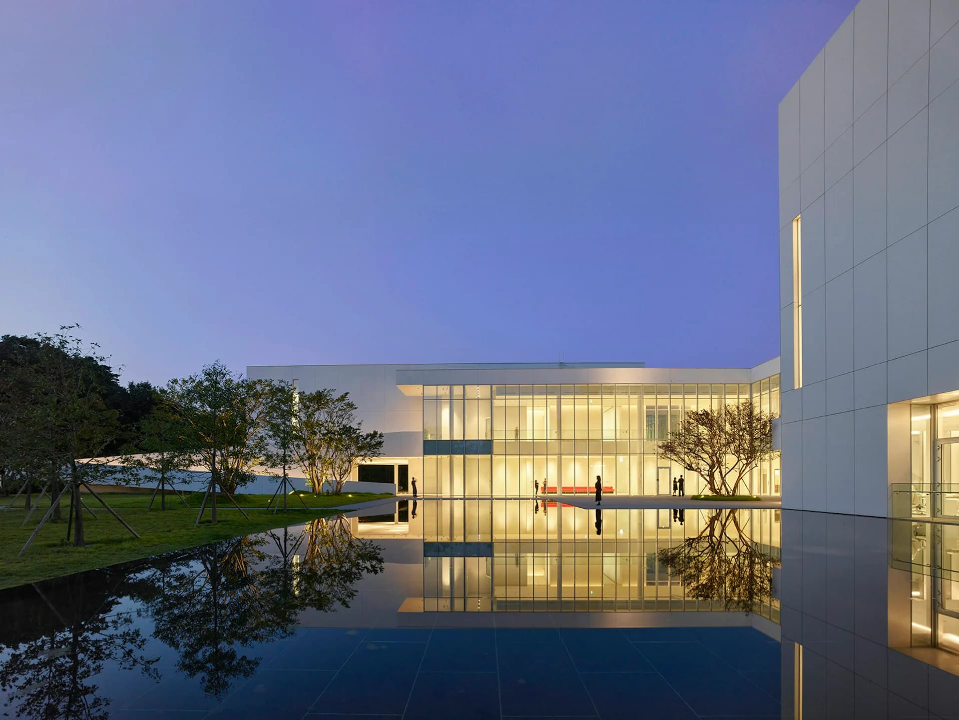 Modern institutional building with glass façade reflected in still water at dusk, illuminated interiors visible, surrounded by manicured landscape by Meier Architects.