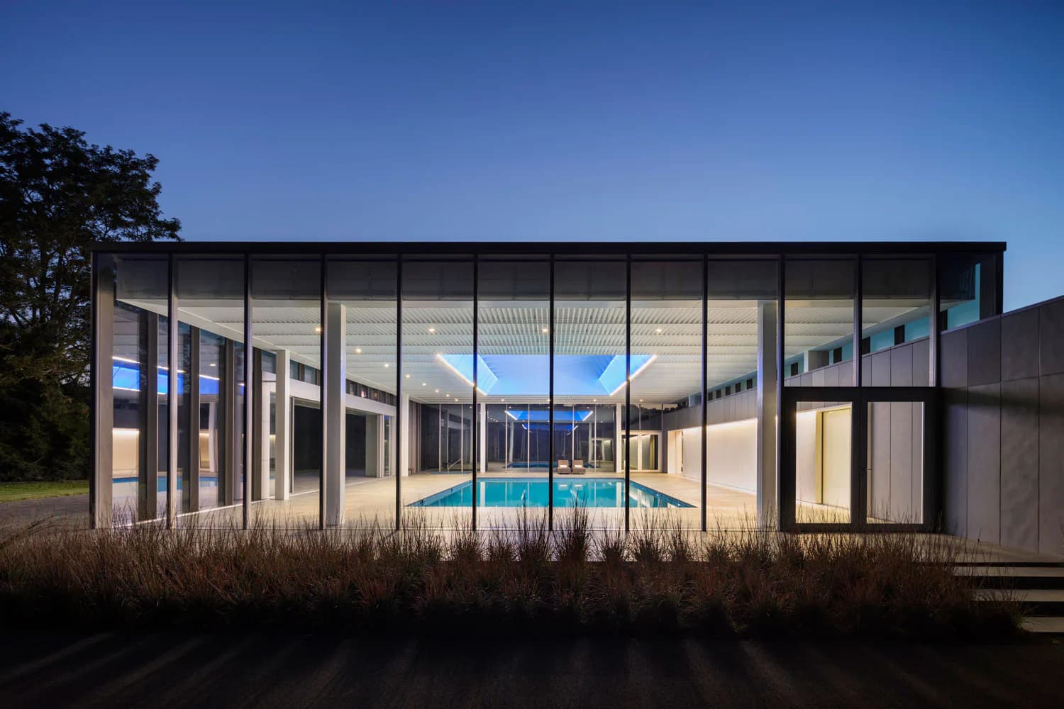 Modern residential architecture with extensive glazing and interior pool visible at dusk, featuring sophisticated lighting design by Melanie Freundlich Lighting Design.