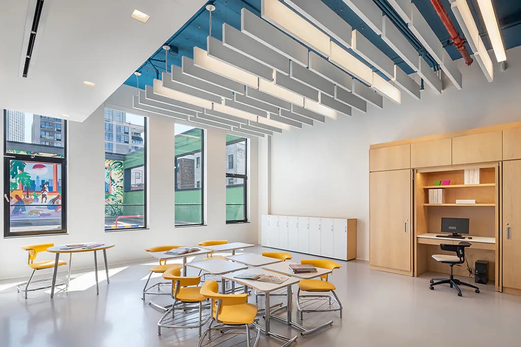 Bright educational classroom with angular linear ceiling lights, yellow chairs, wooden storage cabinetry, and natural daylight from large windows, designed by Melanie Freundlich Lighting Design.