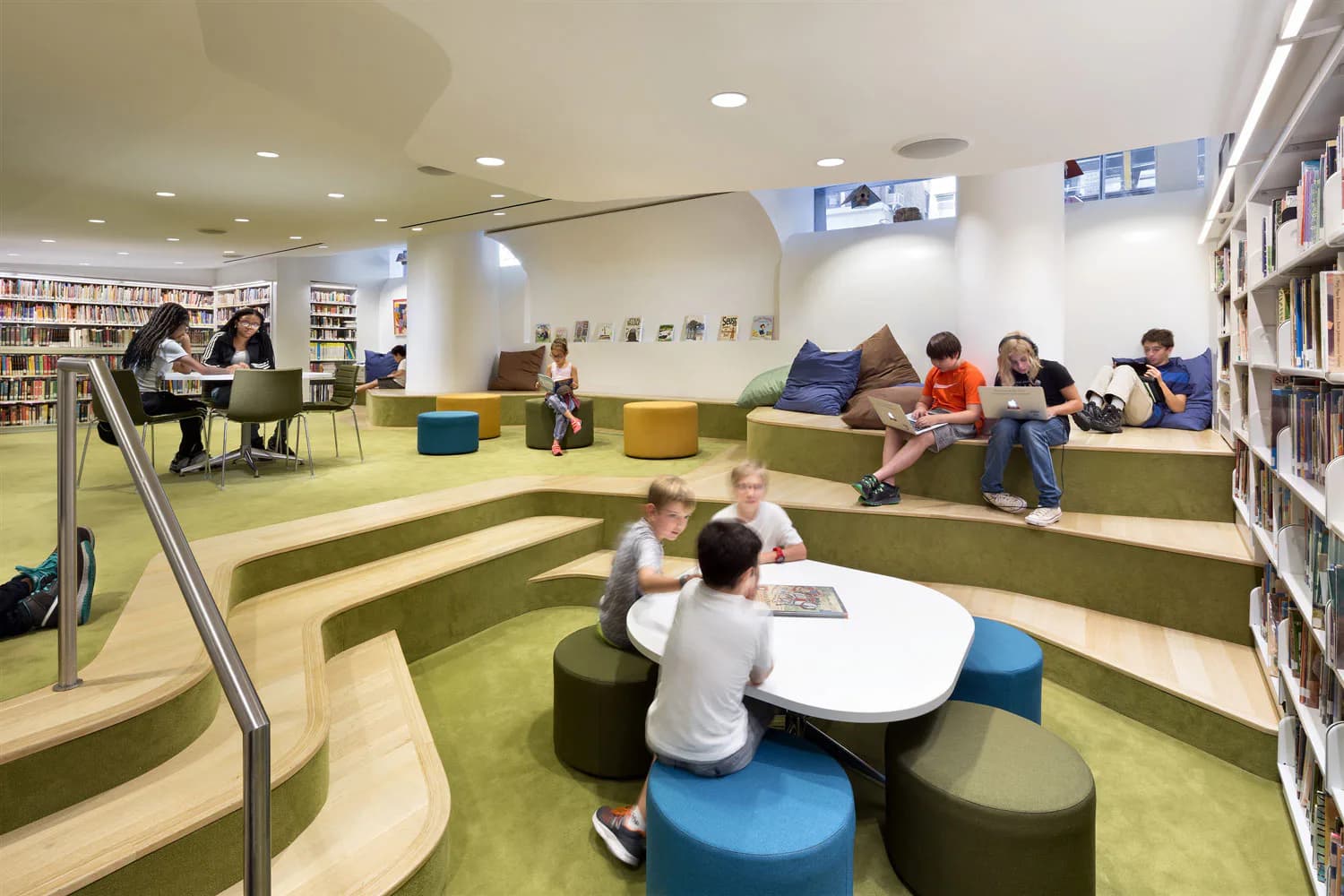Modern library interior with green carpeted tiered seating, blue and olive poufs, bookshelves, and warm ambient lighting design by Melanie Freundlich Lighting Design.