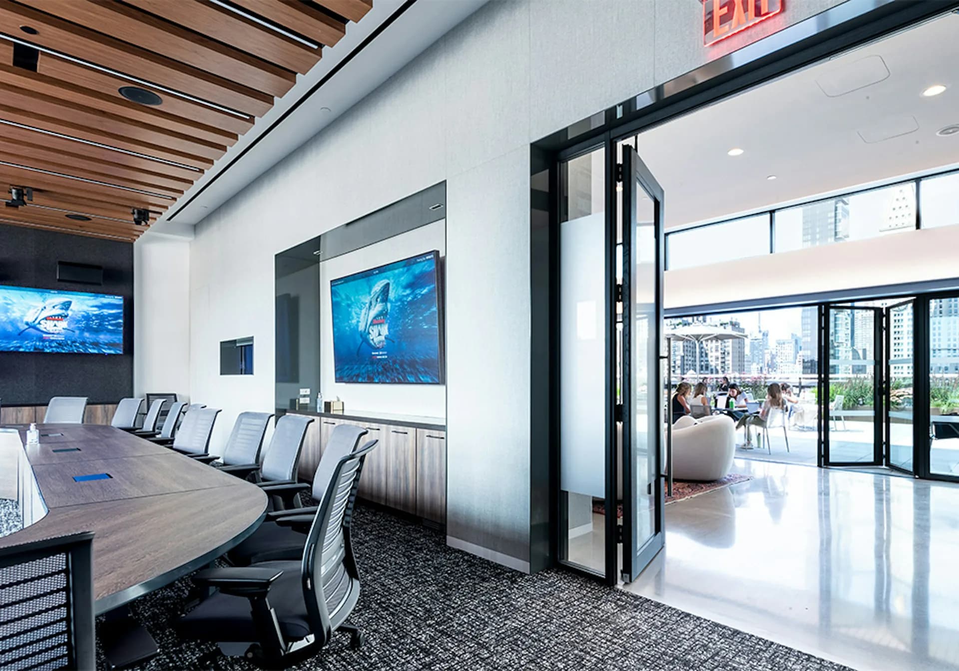 Modern conference room with wooden ceiling, long dark table, ergonomic seating, blue digital displays, and glass doors opening to bright atrium space by Meridian Design Associates, Architects, P.C.