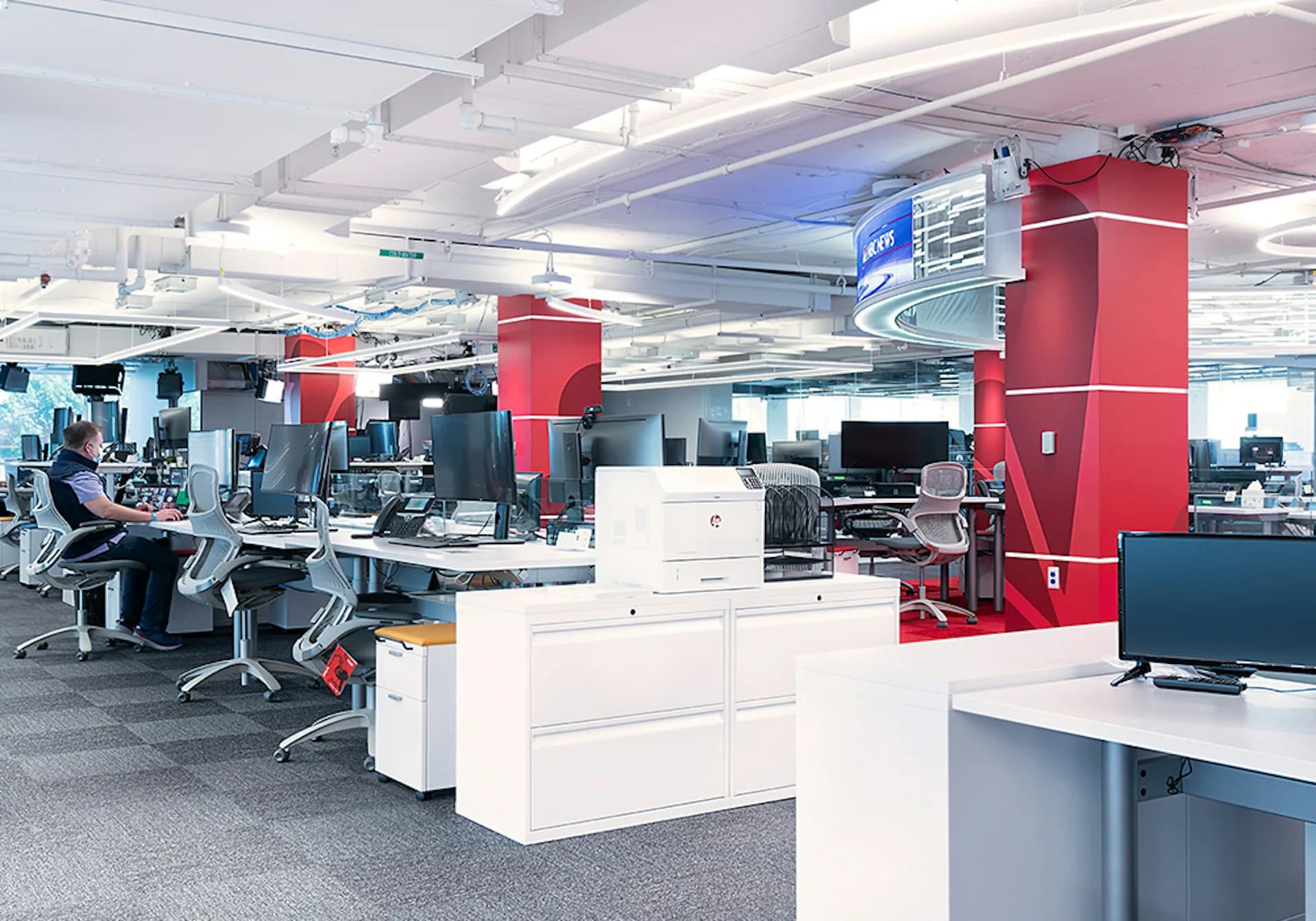 Modern office workspace featuring red structural columns, white workstations, and collaborative areas with blue accent lighting designed by Meridian Design Associates, Architects, P.C.