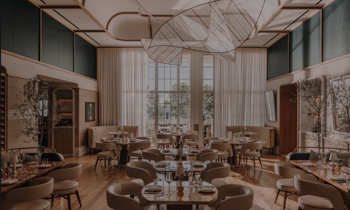 Luxurious restaurant interior with curved banquette seating, floor-to-ceiling windows, geometric ceiling details, and warm wood tones designed by Meyer Davis.