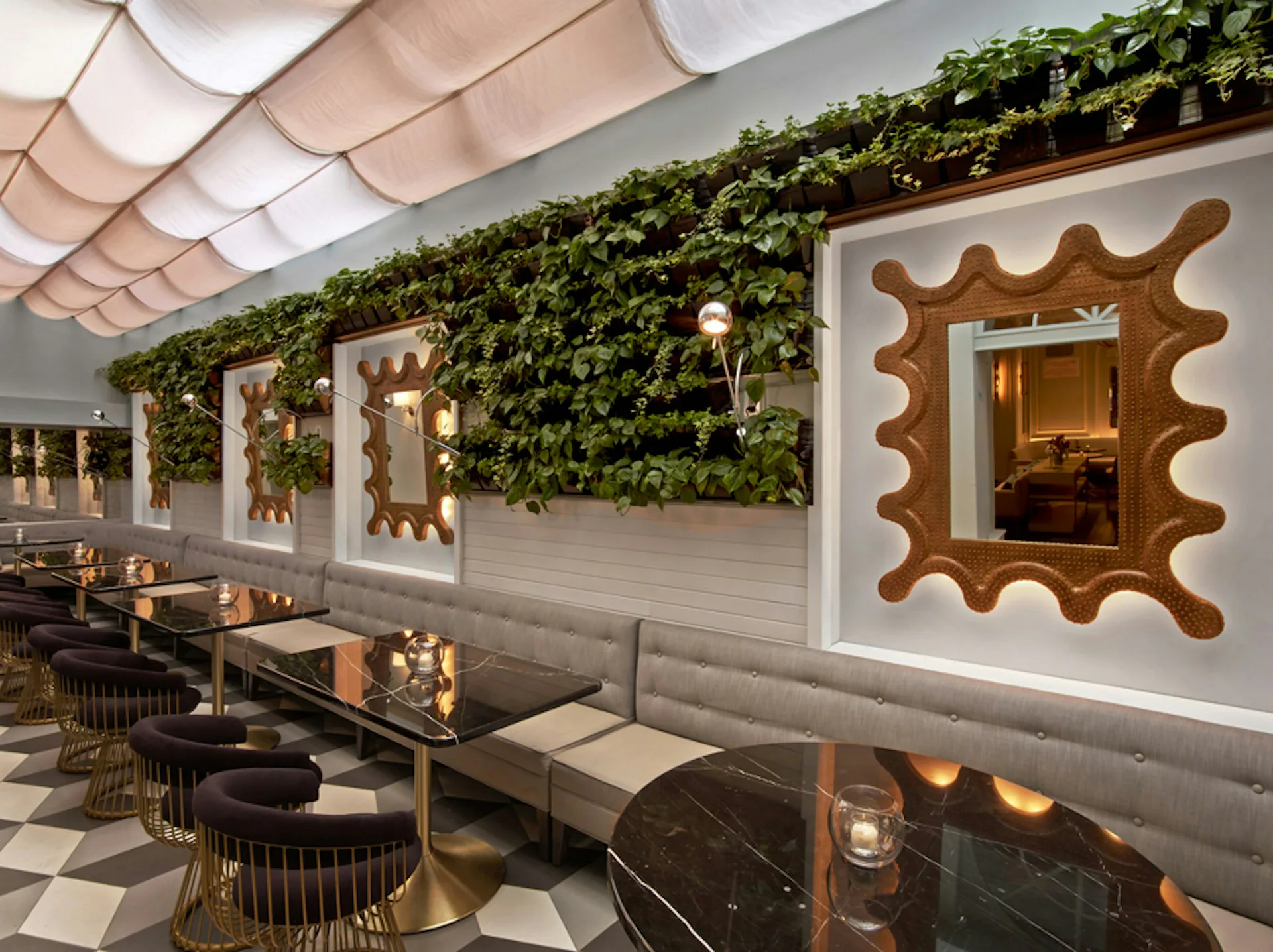 Upscale restaurant interior with gray banquette seating, black tables, decorative wavy mirrors on green ivy-covered wall, and fabric ceiling by Meyer Davis Studio Inc.